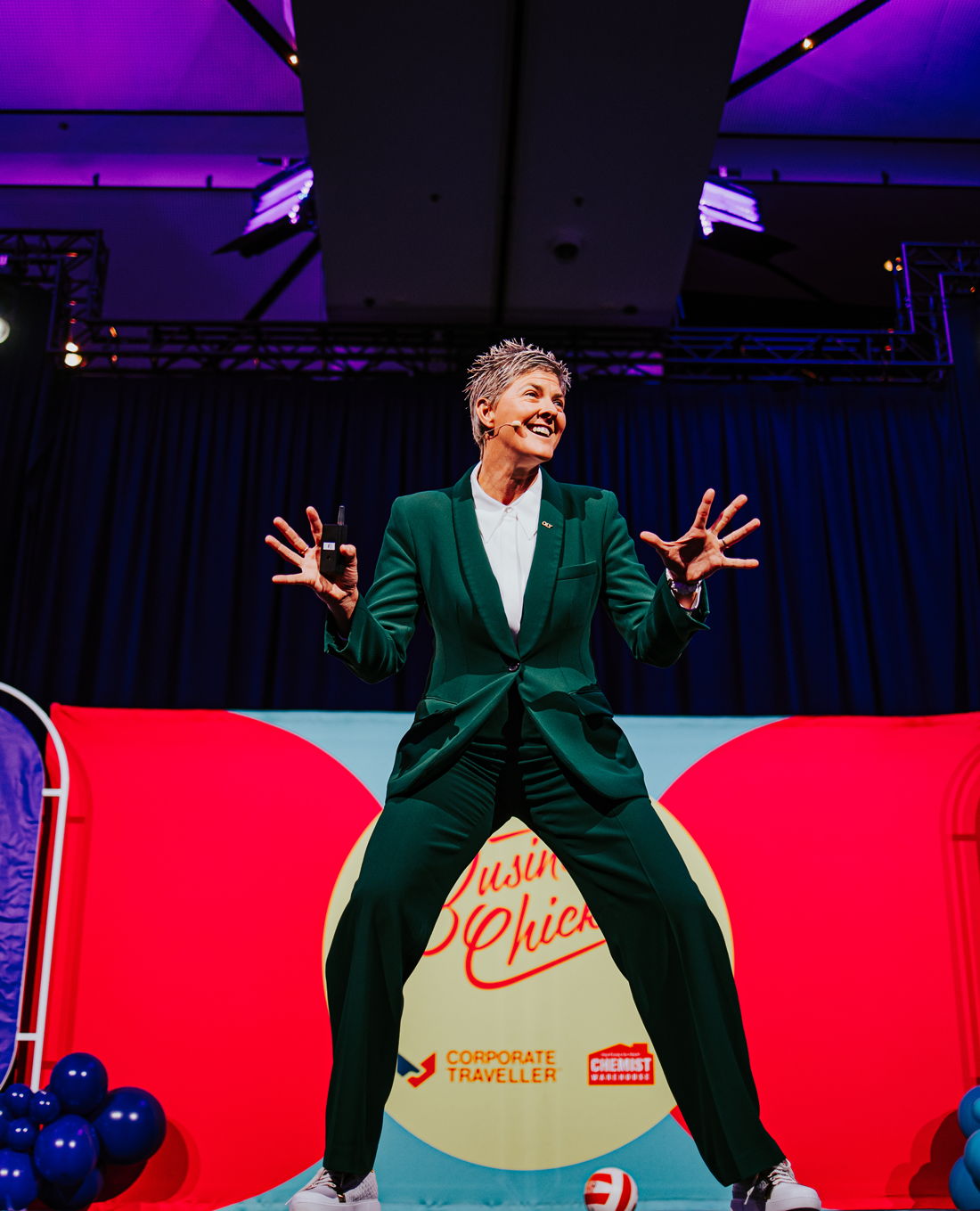 Nat Cook OAM OLY smiling and with her hands extended in a dynamic pose, and wearing a green suit with a white collared shirt and white sneakers.