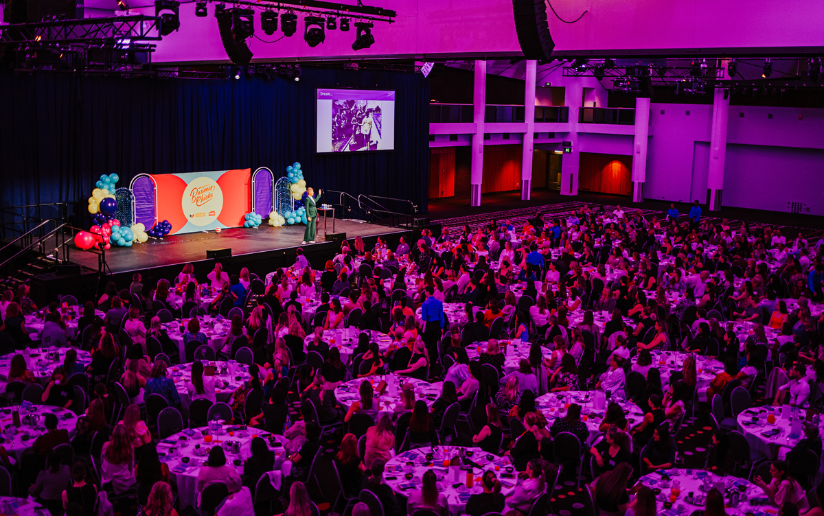 Full room shot showing guests watching Nat Cook OAM OLY speaking on stage, with the room lit in purple