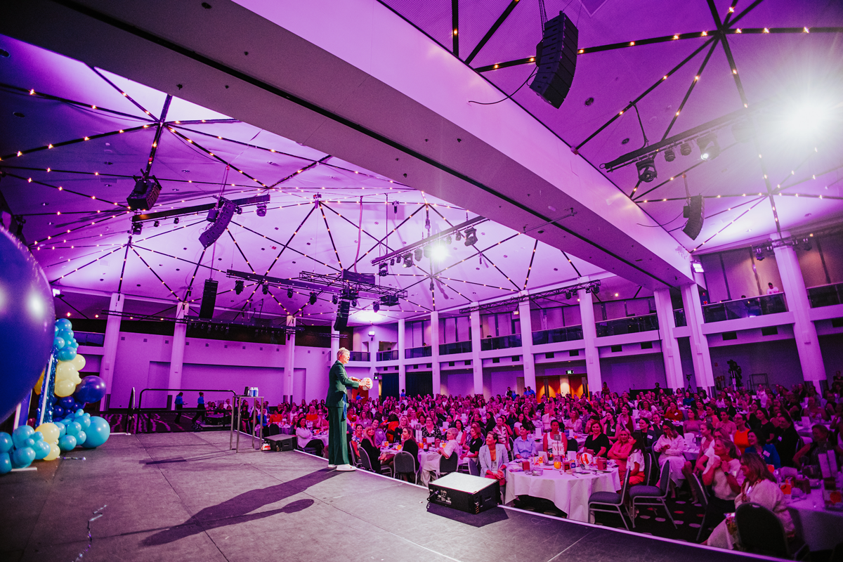 Nat Cook OAM OLY stands on stage facing a crowd, shot from behind, with the room lit up in purple