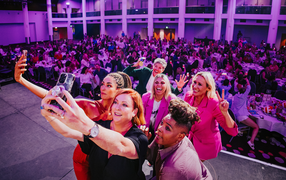 All speakers at the Business Chicks Brisbane event taking selfies on stage, with the audience lit up in purple behind them.