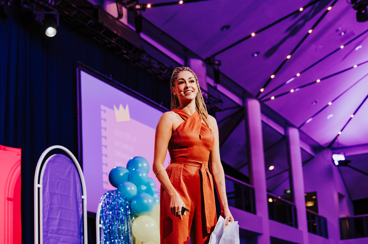 Dr Gina Cleo standing on stage wear a rust coloured jumpsuit. The background of the stage and lights is purple.