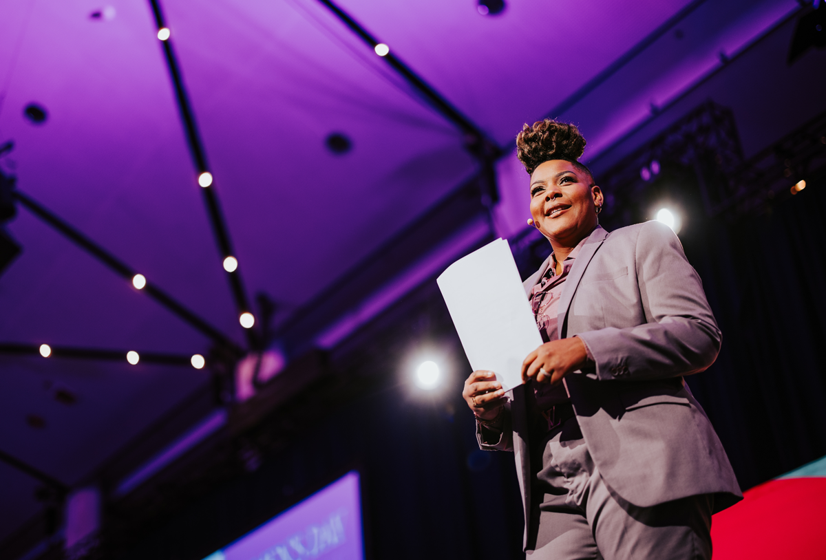 Azure Antoinette smiles on a stage with purple lighting, wearing a grey suit and holding a piece of paper.