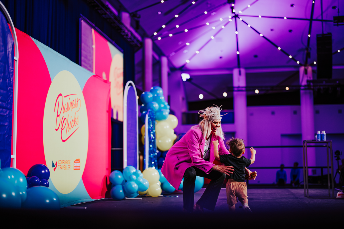 Nartarsha Bamblett is picking up her toddler while she crouches down on stage. Nartarsha is wearing a bright pink blazer and feather headdress.