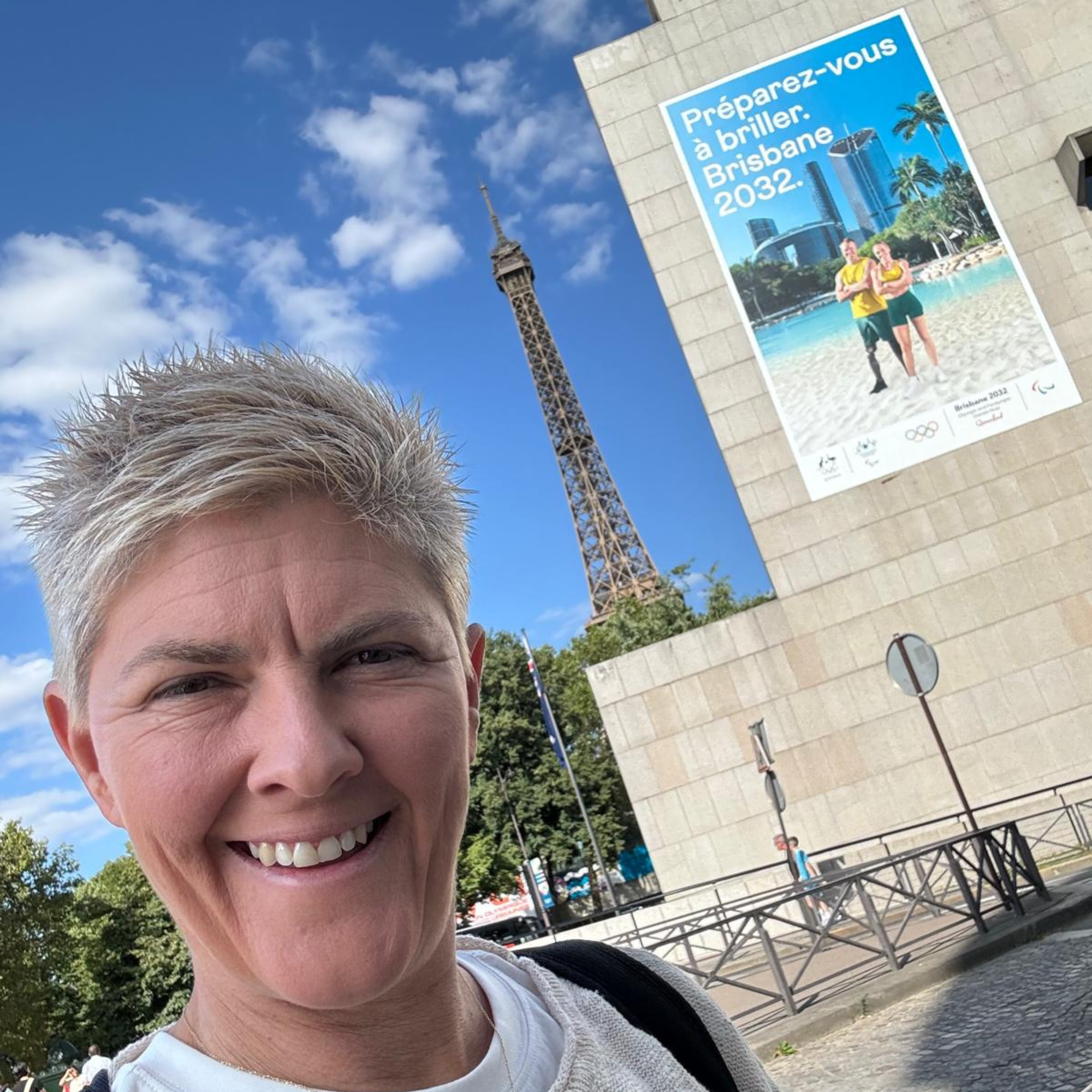 Nat Cook OAM OLY taking a selfie in front of the Eiffel Tower at the Paris 2024 Paralympics welcome event. Nat is standing in front of a billboard for the Brisbane 2032 Paralympic Games