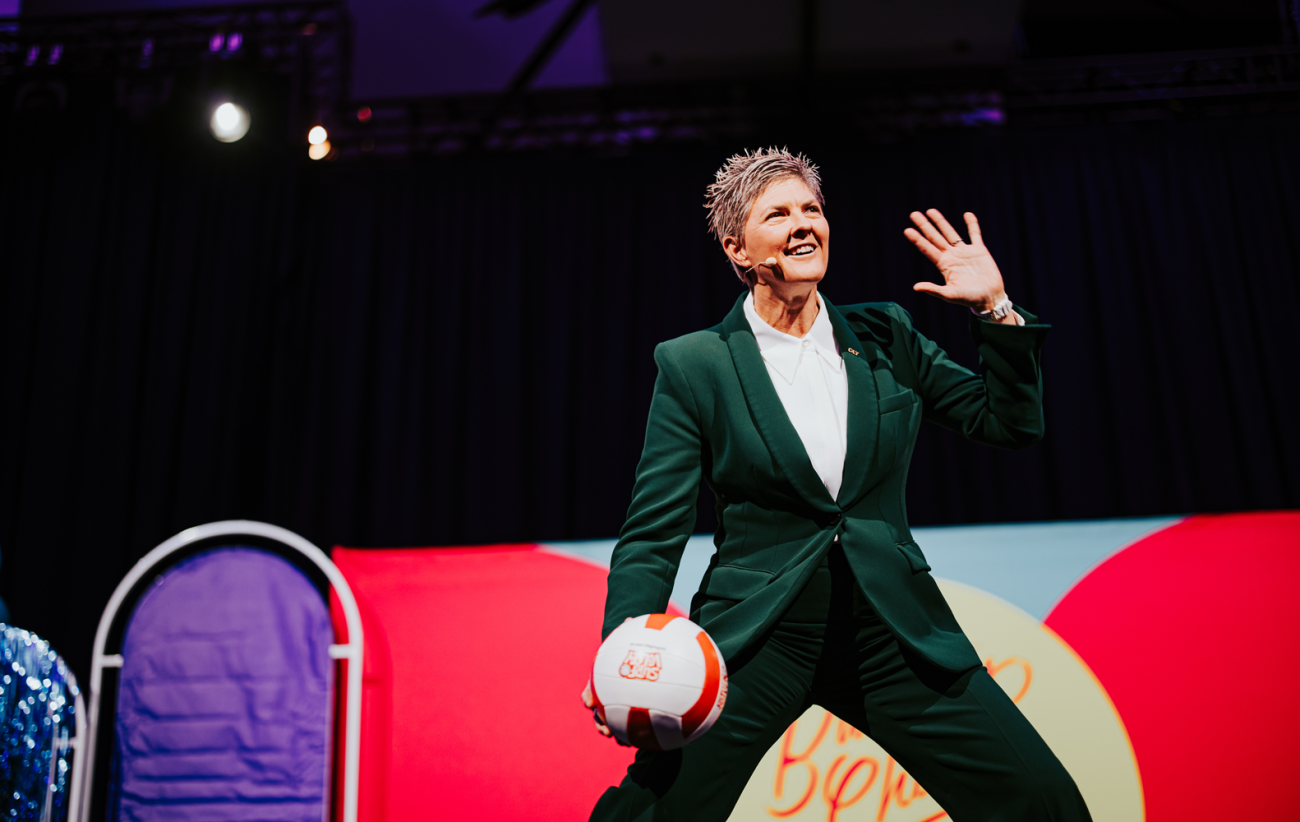 Nat Cook OAM OLY standing on stage holding a volleyball, wearing a green coloured suit and white collared shirt.