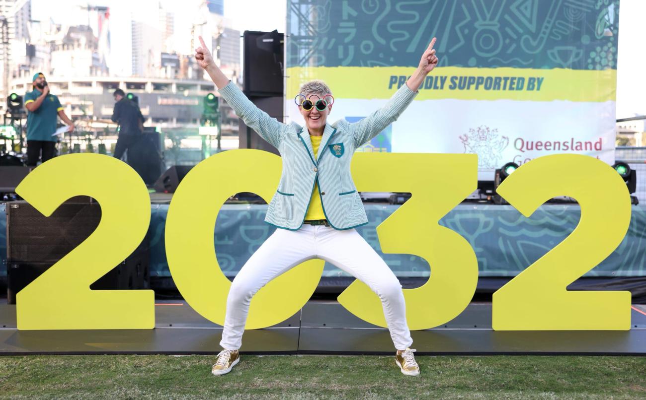 Nat Cook OAM OLY jumping in front of and oversized '2032'. Nat is wearing sunglasses featuring the Olympic rings, and she is wearing her 2000 Australian Olympic team uniform blazer 