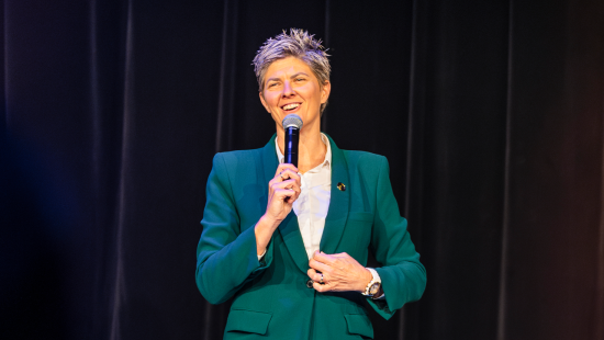 Nat Cook OAM OLY wearing a green suit, standing on stage, smiling and holding a microphone as she addresses an audience (not seen)